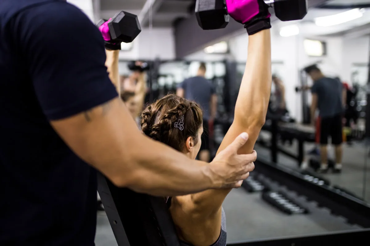 Female client performing shoulder press with personal trainer guidance during private fitness session