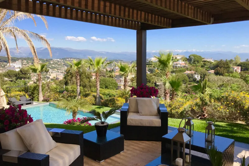villa-gabriela-cannes-rentals-french-riviera-exterior-terrace Villa Gabriela in Cannes – Covered terrace with elegant seating and panoramic views over the French Riviera, palm trees, and a private pool.