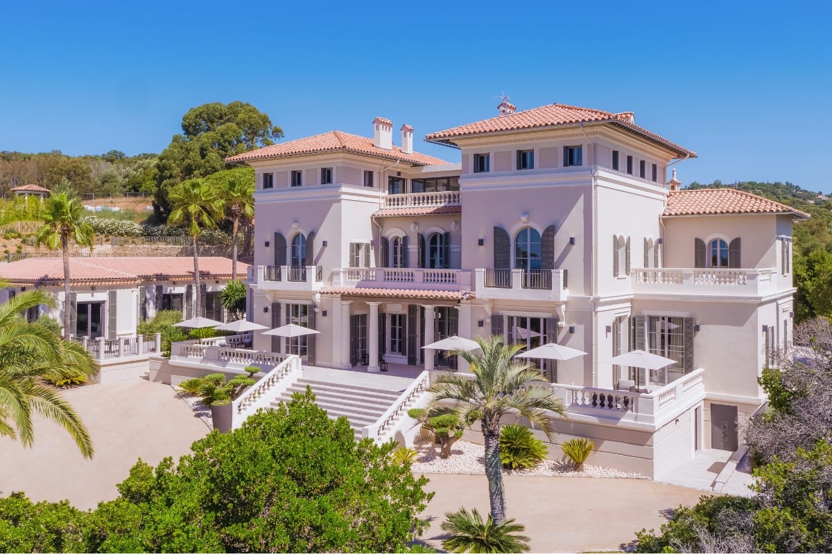 Elegant Mediterranean villa with symmetrical facade, terraces, and palm trees under a clear blue sky.