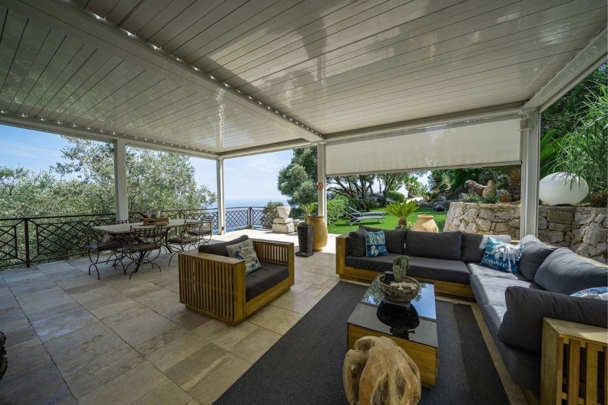 Covered outdoor lounge on a terrace with gray sectional seating, wooden armchairs, coffee table, and dining area; ocean view