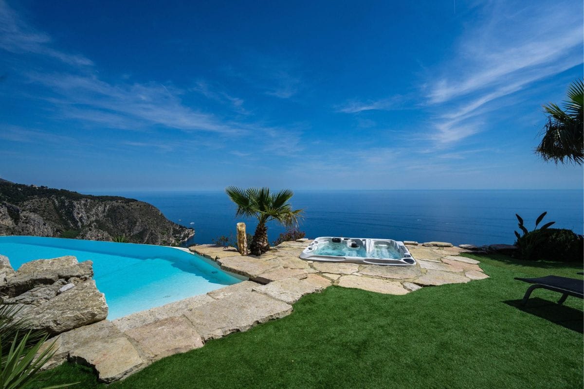 Villa with an infinity pool and a hot tub on a stone terrace overlooking the sea and cliffs, bright blue sky.