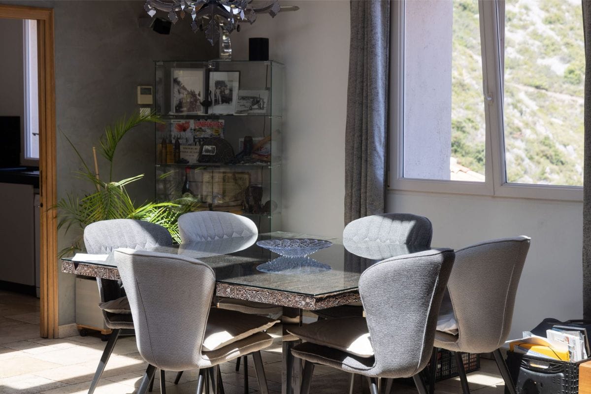 Dining area with a glass-top table surrounded by six light gray upholstered chairs, french doors to the left, large window