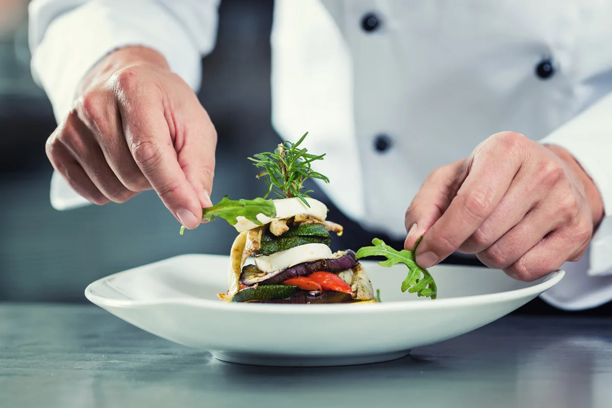 A chef decorates a plated stack of vegetables and greens on a white dish.