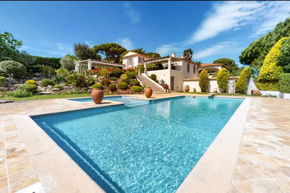 A luxurious villa with a large rectangular swimming pool surrounded by a stone patio and manicured garden plants.