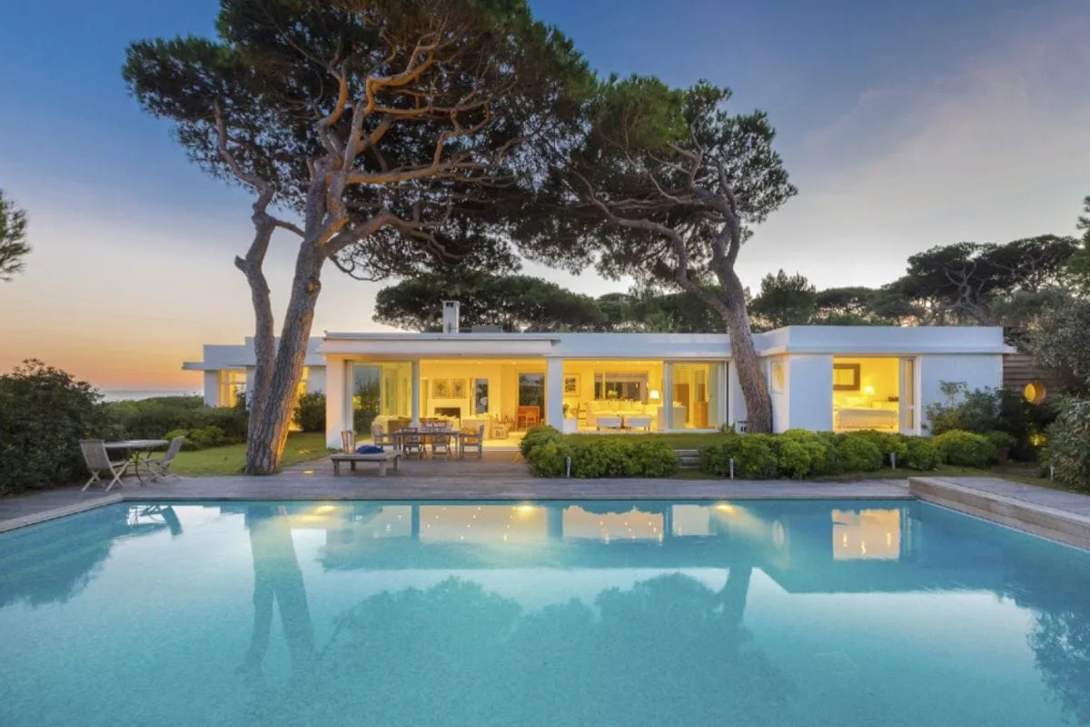 A modern single-story villa with large sliding doors opens to a pool area, surrounded by trees and outdoor seating at dusk.