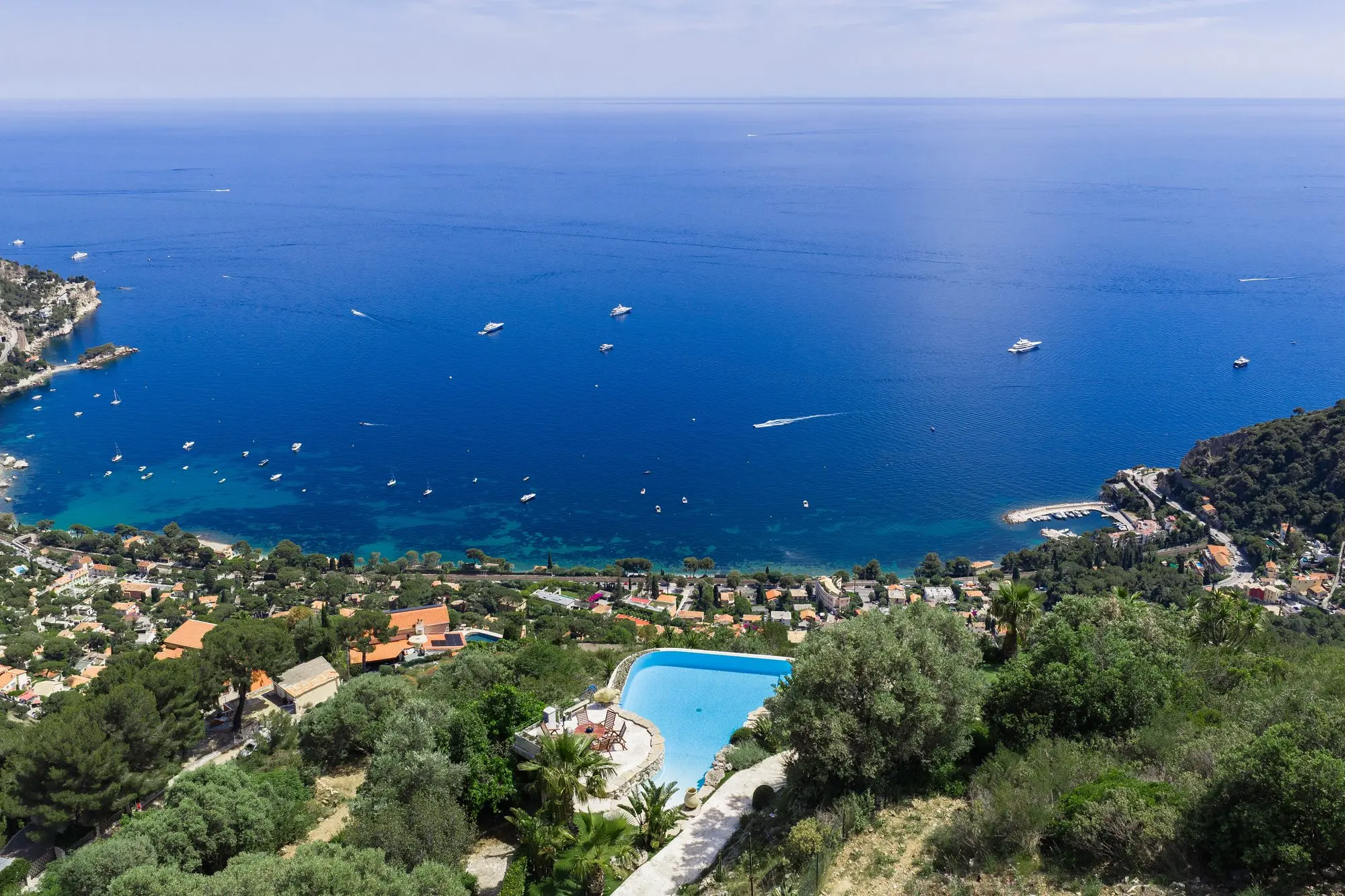 Stunning coastal view of a blue sea with boats, a hillside village below, and a swimming pool nestled among trees in a sunny